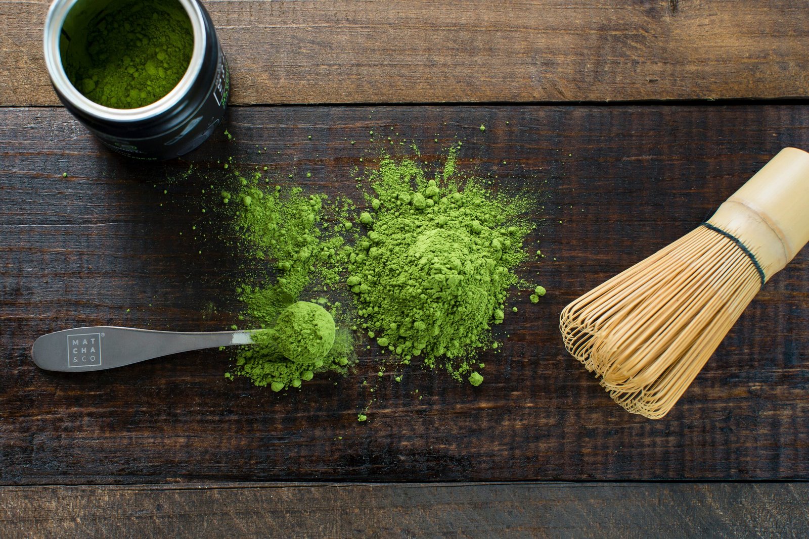 Matcha powder with a bamboo whisk and measuring spoon on a wooden surface.